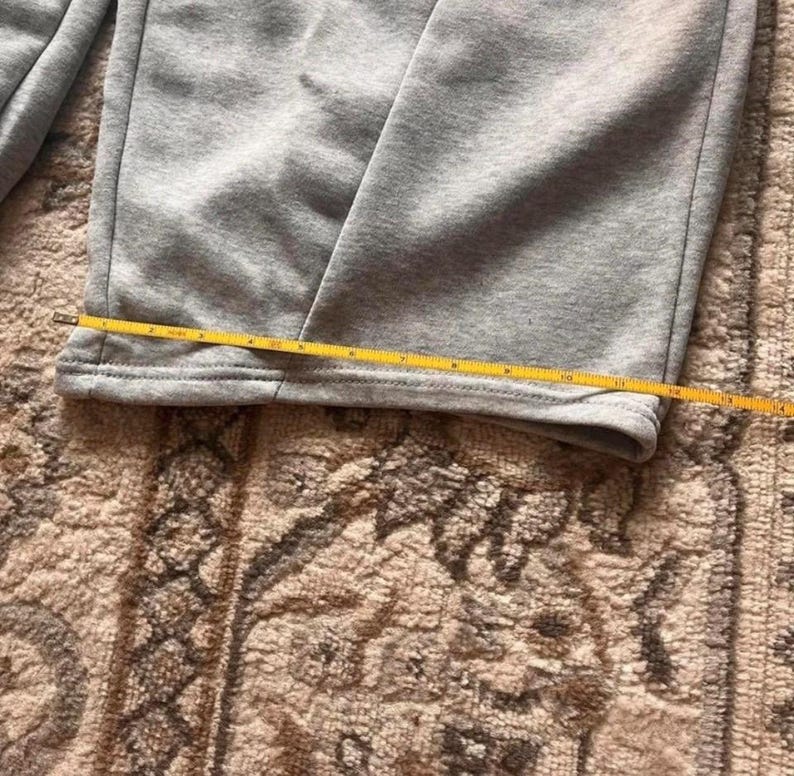 Peut inclure: Un pantalon de jogging gris clair avec une coupe droite et un ourlet cousu. Un m&egrave;tre ruban jaune est plac&eacute; horizontalement sur le bas de la jambe, indiquant une largeur d'environ 26,7 centim&egrave;tres &agrave; cet endroit pr&eacute;cis du v&ecirc;tement.