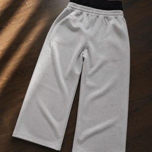 Puede incluir: Un pantal&oacute;n de ch&aacute;ndal gris claro de pernera ancha que presenta una cintura el&aacute;stica de tiro alto con un forro interior negro visible. La prenda est&aacute; confeccionada con un tejido suave y jaspeado e incluye un dobladillo cosido en cada pierna.