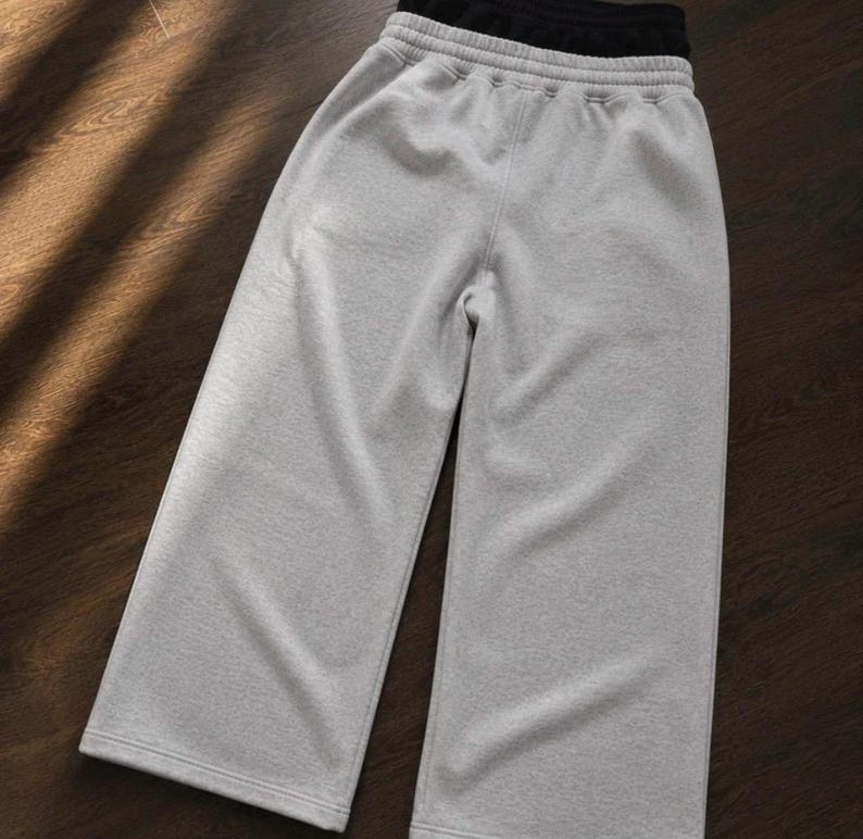 Puede incluir: Un pantal&oacute;n de ch&aacute;ndal gris claro de pernera ancha que presenta una cintura el&aacute;stica de tiro alto con un forro interior negro visible. La prenda est&aacute; confeccionada con un tejido suave y jaspeado e incluye un dobladillo cosido en cada pierna.