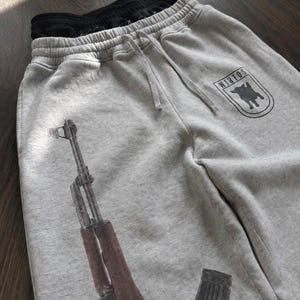 Puede incluir: Pantalones de ch&aacute;ndal de color gris jaspeado claro con un gr&aacute;fico negro de un rifle y un emblema en forma de escudo con el texto WINTOS y la silueta de un perro. Los pantalones tienen una cintura el&aacute;stica con cord&oacute;n y forro negro.