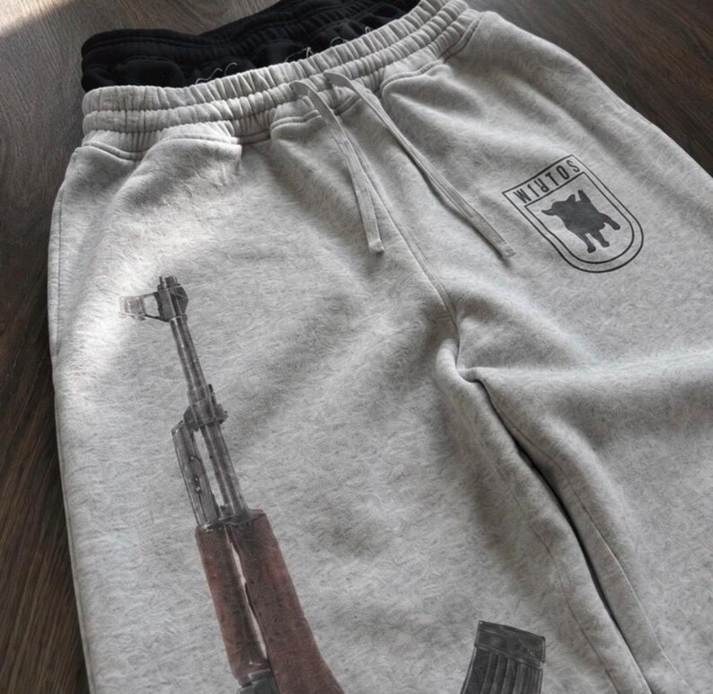 Puede incluir: Pantalones de ch&aacute;ndal de color gris jaspeado claro con un gr&aacute;fico negro de un rifle y un emblema en forma de escudo con el texto WINTOS y la silueta de un perro. Los pantalones tienen una cintura el&aacute;stica con cord&oacute;n y forro negro.