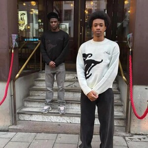 May include: Two individuals wearing long-sleeved sweaters and pants. One wears a black sweater with grey jeans and a black cap. The other wears a white knit sweater with a black graphic design and the word Amici, paired with dark trousers.