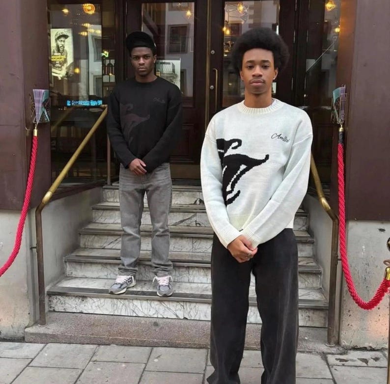 May include: Two individuals wearing long-sleeved sweaters and pants. One wears a black sweater with grey jeans and a black cap. The other wears a white knit sweater with a black graphic design and the word Amici, paired with dark trousers.