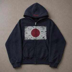 May include: A navy blue zip-up hoodie featuring a distressed graphic of the Japanese flag on the chest. The hood displays the red embroidered text "Synaforce" at the base. The garment includes long sleeves, a front zip fastening, and two pockets.