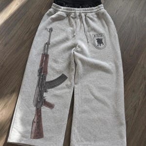 Puede incluir: Pantalones de ch&aacute;ndal de color gris jaspeado claro con un gran estampado gr&aacute;fico de un rifle en la pierna izquierda y un peque&ntilde;o emblema negro en forma de escudo en la pierna derecha con la silueta de una cabra y el texto "WINTOS".