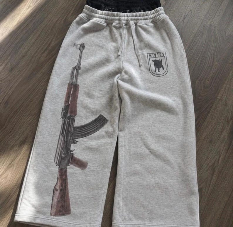 Puede incluir: Pantalones de ch&aacute;ndal de color gris jaspeado claro con un gran estampado gr&aacute;fico de un rifle en la pierna izquierda y un peque&ntilde;o emblema negro en forma de escudo en la pierna derecha con la silueta de una cabra y el texto "WINTOS".