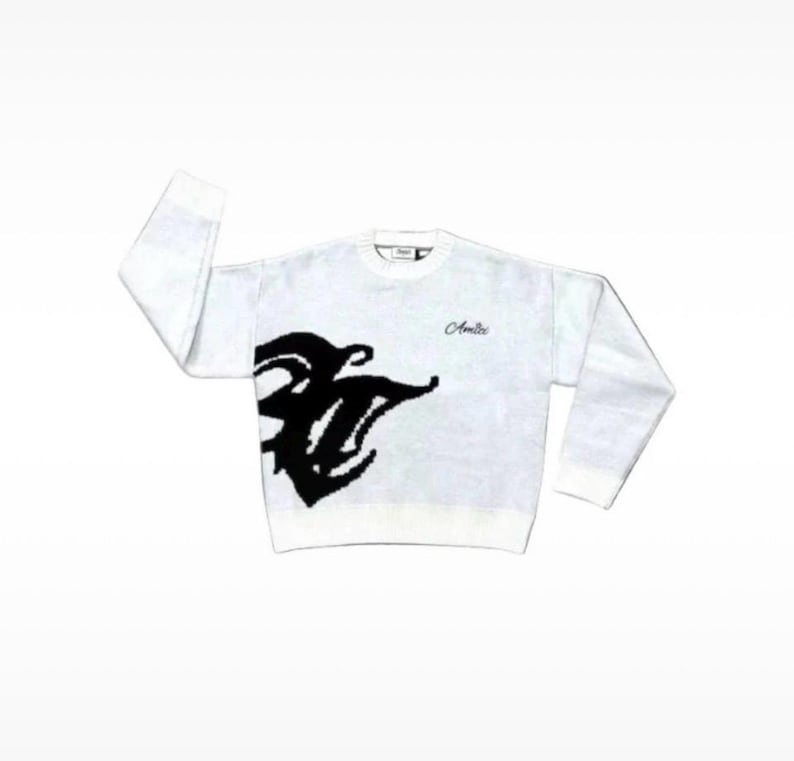 May include: A white long-sleeve crewneck sweater featuring a large black abstract graphic on the lower left side and the word Amiri embroidered in a cursive script on the upper right chest. The garment has a ribbed collar, cuffs, and hem.