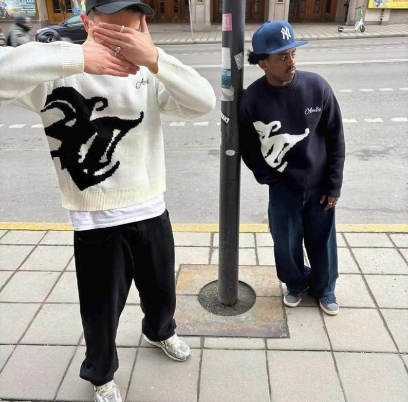 May include: A white long-sleeve knit sweater with a large black graphic design and the text "Amis" on the chest, paired with black trousers. Beside it, a navy blue long-sleeve knit sweater with a white graphic design and "Amis" text, worn with blue jeans.