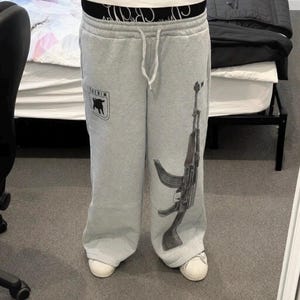 Puede incluir: Pantalones de ch&aacute;ndal gris claro con un gr&aacute;fico negro de un rifle de asalto en la pierna izquierda y un peque&ntilde;o logotipo negro en forma de escudo con la silueta de una cabra en la derecha. Tienen cintura con cord&oacute;n y una banda el&aacute;stica negra.