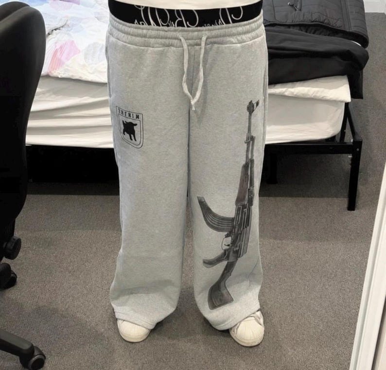 Puede incluir: Pantalones de ch&aacute;ndal gris claro con un gr&aacute;fico negro de un rifle de asalto en la pierna izquierda y un peque&ntilde;o logotipo negro en forma de escudo con la silueta de una cabra en la derecha. Tienen cintura con cord&oacute;n y una banda el&aacute;stica negra.
