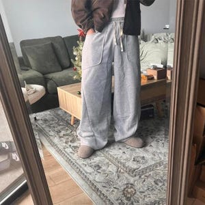 Peut inclure: Pantalon de jogging gris clair &agrave; jambes larges, dot&eacute; d'une taille haute avec cordon de serrage, de deux grandes poches avant aux coutures incurv&eacute;es et d'une coupe ample et d&eacute;contract&eacute;e descendant jusqu'aux chevilles, port&eacute; avec des chaussons beiges.