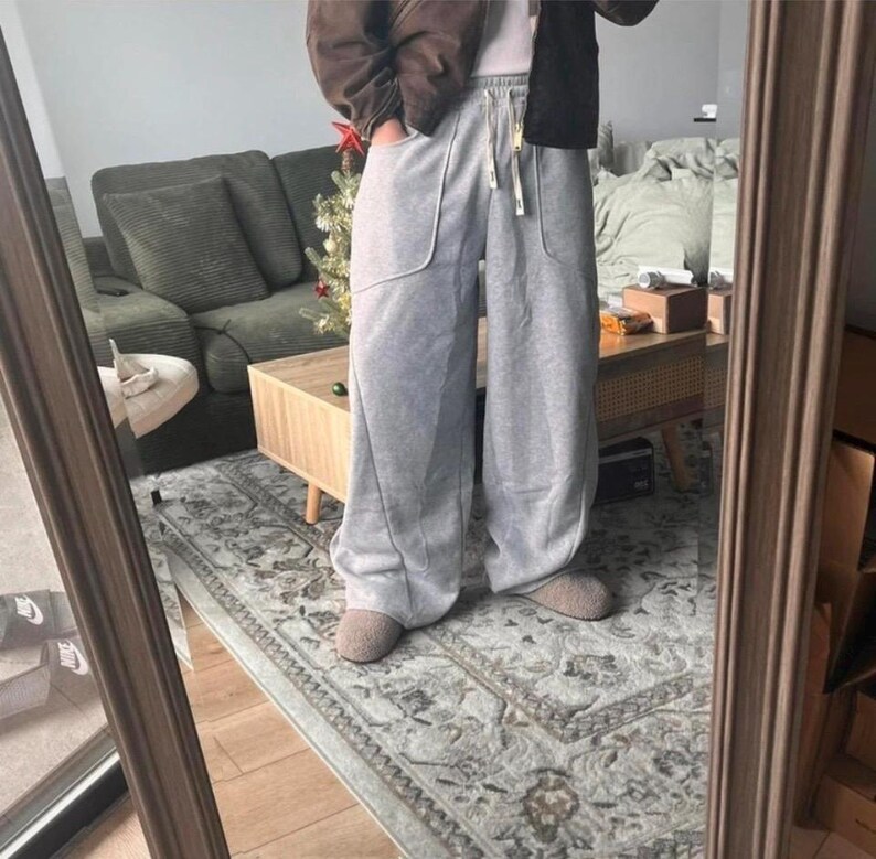 Peut inclure: Pantalon de jogging gris clair &agrave; jambes larges, dot&eacute; d'une taille haute avec cordon de serrage, de deux grandes poches avant aux coutures incurv&eacute;es et d'une coupe ample et d&eacute;contract&eacute;e descendant jusqu'aux chevilles, port&eacute; avec des chaussons beiges.
