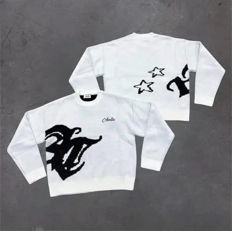 May include: A white long-sleeve crewneck sweater featuring a large black graphic design on the front and back. The front displays the word Amici in cursive script. The back includes two black star outlines and a partial black graphic pattern.