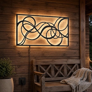 May include: A rectangular wall-mounted light fixture featuring a black metal frame with an intricate, swirling, abstract wire design. The fixture emits a warm, glowing light from behind the metal structure, creating a soft backlight effect.