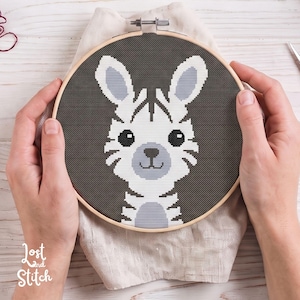 Peut inclure: Un tambour à broder circulaire en bois contenant un motif au point de croix représentant un lapin blanc et gris clair avec de grands yeux noirs sur un fond gris foncé. Le texte Lost and Stitch est imprimé en blanc dans le coin inférieur gauche.