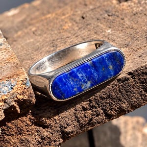 May include: A silver ring featuring a horizontal, elongated oval inlay of deep blue lapis lazuli stone with visible gold-colored pyrite flecks. The metal band has a smooth, polished finish and a thin, beaded texture bordering the stone inlay.