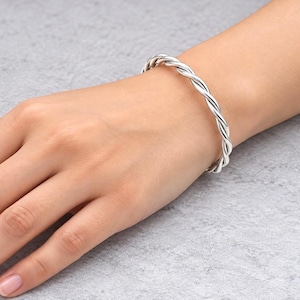 Può includere: Un bracciale rigido color argento lucido caratterizzato da un design a corda intrecciata. Il bracciale è composto da due fili metallici intrecciati che formano una forma circolare continua, creando una superficie liscia e riflettente.