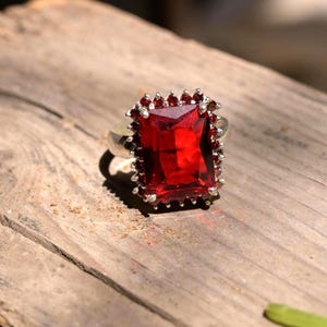 May include: A silver ring featuring a large, rectangular, faceted deep red gemstone as the centrepiece. The main stone is surrounded by a halo of numerous small, round, faceted red stones set in a silver frame with a polished metal band.