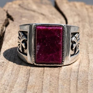 May include: A silver-toned ring featuring a large, rectangular, deep magenta stone set in the center. The band is wide and polished, with detailed, raised cross-like motifs on both sides of the central stone setting, creating a bold appearance.