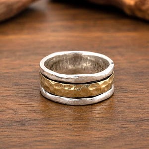 Op de afbeelding: Een brede ring met een centrale gehamerde goudkleurige band, geflankeerd door twee zilverkleurige banden met een onregelmatige, getextureerde afwerking. Het ontwerp bestaat uit drie afzonderlijke lagen die een gelaagd uiterlijk met contrasterende kleuren creëren.