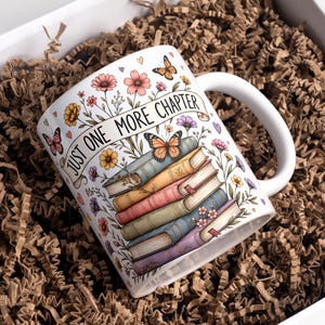 Op de afbeelding: Een witte keramische mok met een kleurrijke illustratie van een stapel boeken, vlinders en diverse bloemen. De tekst "JUST ONE MORE CHAPTER" staat op een banner die over de boeken gedrapeerd is. De mok heeft een gebogen handvat aan de rechterkant.