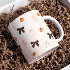 Puede incluir: Una taza de cerámica blanca con un patrón repetitivo de calabazas naranjas, lazos de color marrón oscuro y lazos de cinta de color melocotón claro sobre un fondo de rayas verticales finas de color melocotón claro.