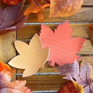 Peut inclure: Deux piles de blocs-notes en papier découpés en forme de feuilles d'érable. Une pile est orange clair et l'autre est d'un orange rougeâtre plus foncé. Les deux présentent de fines lignes blanches horizontales imprimées pour écrire des notes.