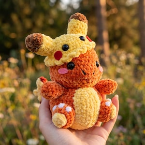 Peut inclure: Une peluche au crochet représentant une petite créature orange au ventre jaune clair, portant un chapeau jaune et marron sur le thème de Pikachu. Le jouet présente des yeux en plastique noir, des joues en feutrine rose et des détails blancs.