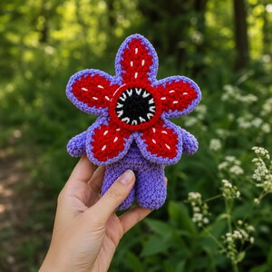 Peut inclure: Une peluche au crochet en forme de créature avec un corps violet et une grande tête en forme de fleur à cinq pétales. Les pétales sont rouges avec des accents blancs brodés, entourant un centre circulaire noir avec des dents blanches pointues.