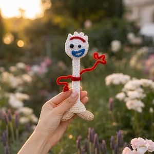 Peut inclure: Une figurine jouet crochetée ressemblant à un personnage spork avec une tête et un corps blancs, des bras en cure-pipe rouge, une bouche en laine bleue et deux yeux mobiles dépareillés. La figurine a des pieds beiges et des mailles détaillées.