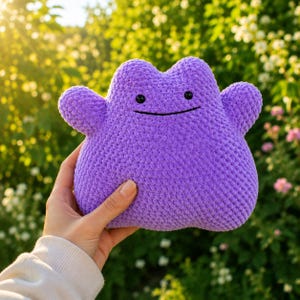May include: A purple crochet plush toy shaped like a blob with two small arms, a rounded top, and a simple face featuring two small black circular eyes and a thin, horizontal black line for a mouth, crafted with a textured yarn stitch pattern.