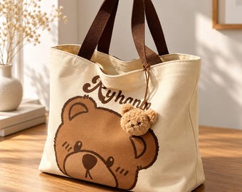 May include: A cream-colored canvas tote bag featuring a large brown bear face graphic and the text "Ryhanm" printed in brown. The bag has two brown fabric handles and includes a small, fuzzy, light brown plush bear charm attached.