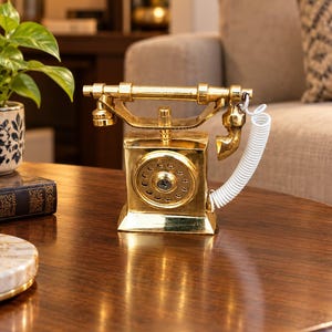 May include: A polished gold-tone metal decorative telephone figurine featuring a classic cradle design, a circular rotary dial face, and a white coiled plastic cord connecting the handset to the base unit, creating a shiny reflective surface.