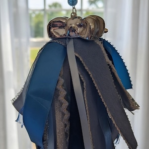 Pode incluir: Um ornamento suspenso com um laço prateado brilhante no topo e um conjunto de fitas de tecido em camadas em tons de azul, cinza e branco. As fitas possuem texturas variadas, incluindo cetim, bordas desfiadas e detalhes com brilho.