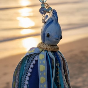 Puede incluir: Un adorno colgante que presenta una cabeza de pez esculpida en azul claro con ojos oscuros, unida a una cinta azul claro. Debajo de la cabeza hay un grupo de varias cintas de tela y adornos en tonos azul, turquesa y amarillo con cuentas.