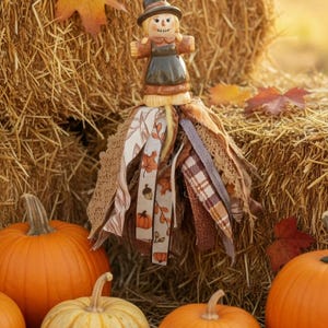 May include: A hanging scarecrow ornament featuring a painted wooden figure with a black hat and brown dress, suspended by a string with green and yellow beads. The base consists of a layered tassel made of various fabric strips in autumn colors.