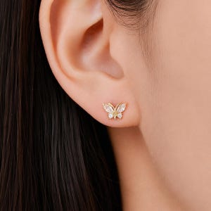 Peut inclure: Une boucle d'oreille puce en forme de papillon dorée, ornée de deux pierres claires taille marquise pour les ailes supérieures et de deux pierres rondes pour les ailes inférieures, avec une petite pierre centrale sur une monture dorée.