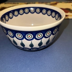 May include: A ceramic bowl with a cream-colored base featuring a hand-painted pattern of dark blue concentric circles with green and orange centers, accented by dark teal leaf-shaped motifs around the lower exterior of the bowl.