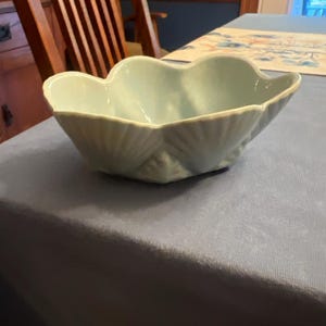 May include: A pale blue ceramic bowl shaped like a large seashell with a scalloped rim and a textured exterior featuring vertical ridges that mimic the natural lines of a shell. The bowl has a smooth, glossy finish and a wide, open basin.