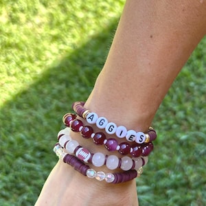 Op de afbeelding: Een stapel van vijf kralenarmbanden met kastanjebruine kleischijfjes, doorschijnende witte kralen en donkerpaarse ronde kralen. Eén armband bevat witte letterkralen die AGGIES spellen en een andere heeft een kleine bedel in de vorm van een football.