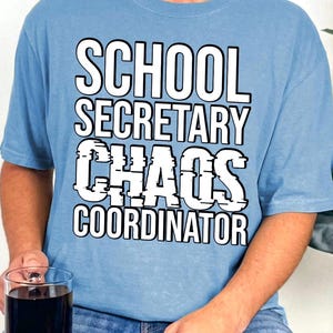 以下が含まれることがあります： 太字のサンセリフ体で「SCHOOL SECRETARY CHAOS COORDINATOR」という白い文字がプリントされたライトブルーのTシャツ。デザインは衣服の胸の中央に配置されており、半袖でクラシックなクルーネックのスタイルです。