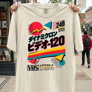 May include: A cream-colored short-sleeve t-shirt featuring a graphic print with the text "246 MINUTES NETTERS", "ダイナミクロン", "ビデオ-120", and "VHS". The design includes geometric shapes, a red circle, and colorful stripes in red, yellow, and teal.