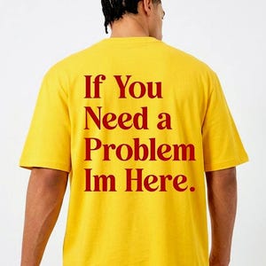 May include: A bright yellow short-sleeved t-shirt featuring red text printed on the back. The text reads, "If You Need a Problem Im Here." The shirt has a crew neckline and a standard fit, showcasing the bold red lettering against the yellow fabric.