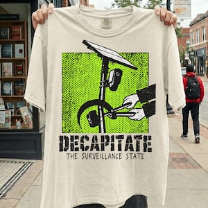 以下が含まれることがあります： クリーム色のTシャツで、監視カメラのポールをボルトカッターで切断する手のグラフィックが描かれています。デザインは明るい緑色のドット柄の正方形の中に配置されています。グラフィックの下には「DECAPITATE THE SURVEILLANCE STATE」という文字が記されています。