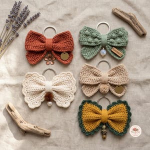 Puede incluir: Cinco llaveros de lazos de ganchillo en colores óxido, verde salvia, crema, beige y amarillo mostaza con borde verde oscuro. Cada lazo incluye una anilla metálica y adornos como cuentas de madera, dijes metálicos o pequeños pompones.