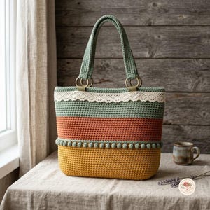 May include: A crochet tote bag featuring horizontal stripes in sage green, terracotta, and mustard yellow. The bag includes a cream-coloured lace trim along the top, a row of small pom-poms above the yellow section, and two sturdy green crochet handles.