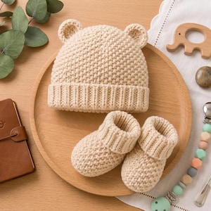 Könnte beinhalten: Eine beige gestrickte Babymütze mit kleinen Bärenohren und ein dazu passendes Paar gestrickter Babyschuhe. Die Mütze hat ein strukturiertes Strickmuster und ein geripptes Bündchen, während die Schuhe ein ähnliches Design mit gerippten Öffnungen aufweisen.
