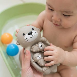 Puede incluir: Un juguete de baño de goma marrón claro con forma de nutria grande que sostiene dos figuras de nutria más pequeñas en su vientre. El juguete tiene una mancha blanca en la cara con ojos negros, nariz negra y bigotes. La superficie está mojada.