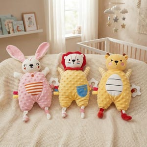 May include: Three plush toys featuring a pink rabbit, a yellow lion, and a yellow bear. Each toy has a textured dimpled body, a front pocket, and small attached fabric tags. The rabbit has long ears, the lion has a red mane, and the bear has ears.