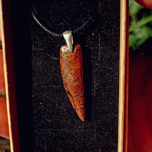 Peut inclure: Un pendentif composé d'une pierre longue et effilée brun rougeâtre avec des éclats irisés verts et bleus, maintenu par une bélière en métal argenté sur un cordon noir fin, présenté dans une boîte en carton rectangulaire avec un insert en mousse noire.